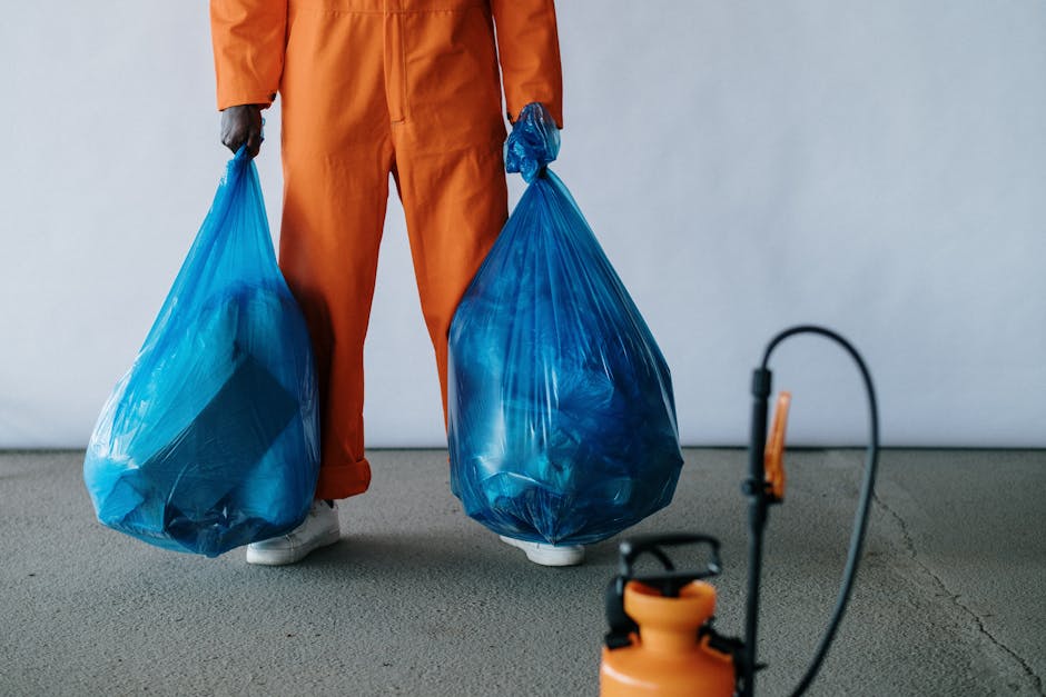 A person wearing orange work overalls and white shoes is standing on a grey concrete floor, holding two large blue plastic rubbish bags filled with waste material. The bags appear full and are tied at the top, with the person gripping each bag’s handles. The background is a plain, light-colored wall, creating a neutral setting typical of an industrial or waste collection environment. To the right front of the person, there is a small orange portable sprayer or disinfectant bottle with a black trigger and a hose, indicating a cleaning or sanitization process often associated with rubbish clearance services. The scene reflects a professional waste removal activity, with clear emphasis on the handling of refuse in a manner consistent with private waste collection or on-site clearance, aligning with services provided by rubbishremovalbrixton.org.uk.