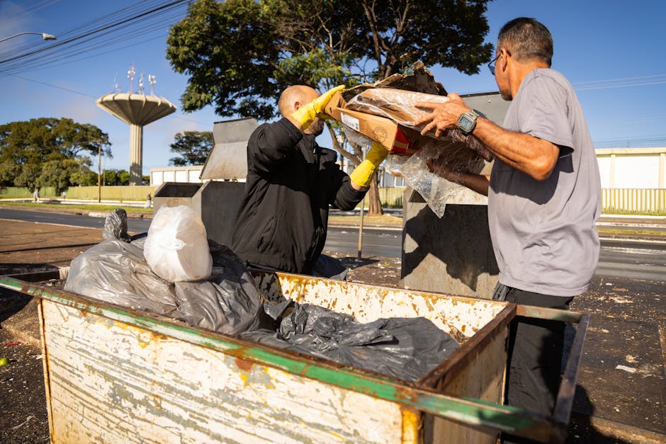 Two men are outdoors, engaged in rubbish collection or disposal activities, with one standing inside a large, open metal container or skip that shows signs of rust and wear. The man on the left, wearing a black jacket and yellow gloves, is placing or lifting a cardboard box filled with mixed debris and possibly construction waste, with some of the contents wrapped in clear plastic. The other man, dressed in a light grey shirt and dark trousers, is helping or receiving the box, with both adjusting or transferring materials between each other. The environment features a paved surface, with a background of trees, a white building with a yellow railing, and a bright blue sky indicating daytime. There are power lines overhead, and the overall scene reflects a practical waste management effort, aligning with private rubbish removal services like those offered by Rubbish Removal Brixton, focusing on efficient on-site clearance of various waste materials.