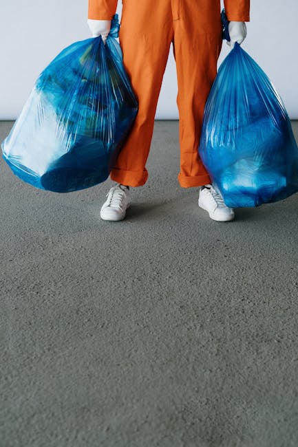 A person wearing orange work overalls and white shoes is standing on a grey concrete floor, holding two large blue plastic rubbish bags filled with waste material. The bags appear full and are tied at the top, with the person gripping each bag’s handles. The background is a plain, light-colored wall, creating a neutral setting typical of an industrial or waste collection environment. To the right front of the person, there is a small orange portable sprayer or disinfectant bottle with a black trigger and a hose, indicating a cleaning or sanitization process often associated with rubbish clearance services. The scene reflects a professional waste removal activity, with clear emphasis on the handling of refuse in a manner consistent with private waste collection or on-site clearance, aligning with services provided by rubbishremovalbrixton.org.uk.