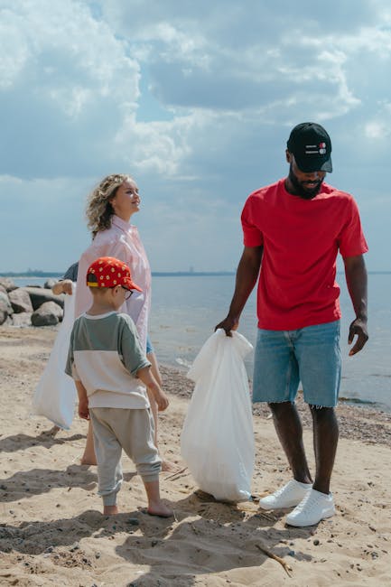 A man with a dark beard, wearing a red t-shirt, light blue denim shorts, white sneakers, and a navy cap with red and white accents, is standing on a sandy beach, holding a large white plastic bag with his right hand. To his left, a young boy with a red cap featuring yellow polka dots, a grey and white t-shirt, and beige shorts, is also holding a plastic bag and facing towards the man. Behind them, an older woman with curly blonde hair, wearing a light pink dress, stands near the water's edge, looking towards the horizon. The background features a cloudy blue sky and calm water with a rocky shoreline in the distance. The scene depicts a clean-up activity at the beach, illustrating waste collection and environmental care, which aligns with local rubbish removal services focused on outdoor and shoreline waste management.