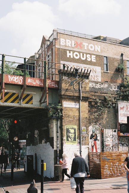 A multi-story brick building with a weathered exterior, featuring large painted signage that spells out 'BRIXTON' in bold white letters with a red 'X' and 'HOUSE' beneath it, partially obscured by graffiti and posters. An elevated railway track runs below the building, with a metal safety railing and a red and yellow striped warning sign visible on the underside. Several posters and graffiti adorn the lower wall, and a drainpipe extends from the roof down along the brickwork. In the foreground, two pedestrians, one in a black jacket and the other in a pink top, walk along the street, with additional people and elements of urban street furniture visible near the footpath. The overall scene portrays an urban setting with an association to local cultural or community activity, typical of an area with independent waste removal services like those provided by Rubbish Removal Brixton, emphasizing the practical aspect of waste management in busy residential and commercial environments.