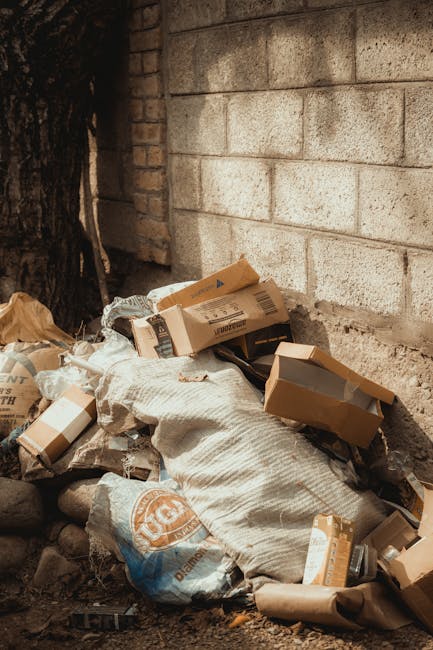 A pile of discarded cardboard boxes, some torn and flattened, stacked against a rough brick wall with visible mortar lines near a concrete or stone wall section. The boxes vary in size and color, primarily brown, with some featuring printed labels and barcodes. Surrounding the boxes are crumpled brown paper and plastic packaging materials, and a large white woven sack partially covering part of the debris. The scene is outdoors, with natural light illuminating the waste and cast shadows suggesting a partly shaded environment. The rubble is placed on uneven ground mixed with small stones and dirt, indicating a waste collection or disposal area. The background includes a darkened section of tree bark or a wooden surface nearby. This setup reflects an off-site or private rubbish removal activity, consistent with independent waste handling efforts by Rubbish Removal Brixton, supporting alternative rubbish collection services in urban environments.