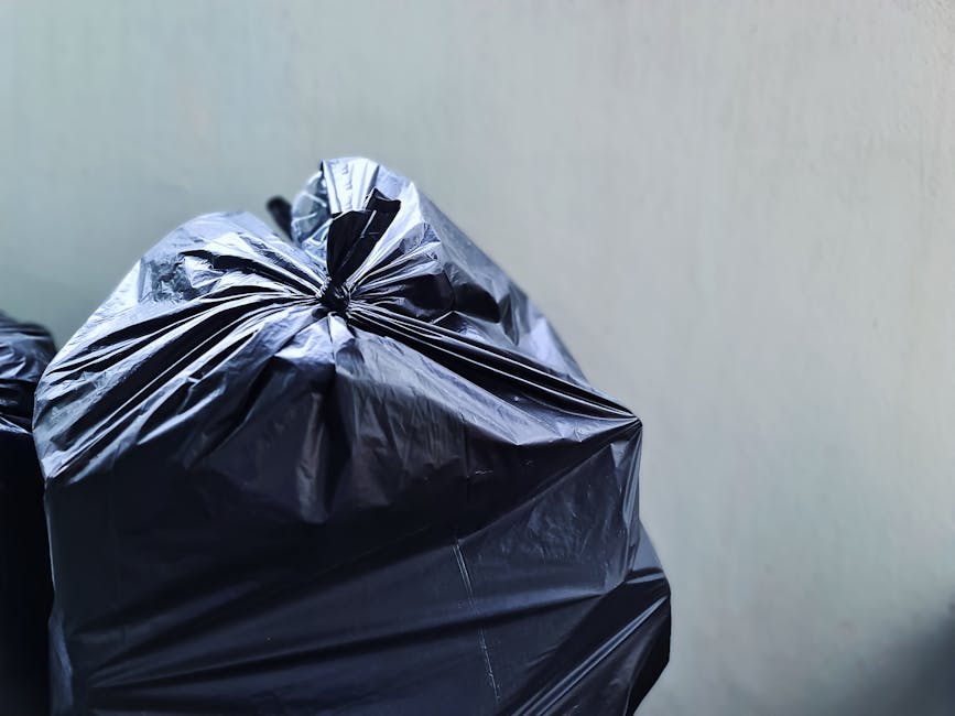 A close-up of a large black rubbish bag made of glossy plastic, tightly knotted at the top, positioned on the left side of the image. The bag appears full and has visible creases and slight folds, indicating it is filled with waste materials. The background features a plain, light-colored wall with a smooth surface. The lighting is soft and even, with no direct shadows, suggesting natural or diffused indoor light. The scene captures a typical setting for private waste collection, such as an on-site rubbish disposal area or a commercial waste handling environment, with the focus on the disposal bag as part of waste removal services like those offered by Rubbish Removal Brixton. The context implies preparing rubbish for collection or disposal in an urban environment, relevant to alternative waste handling methods outside of local authority services.