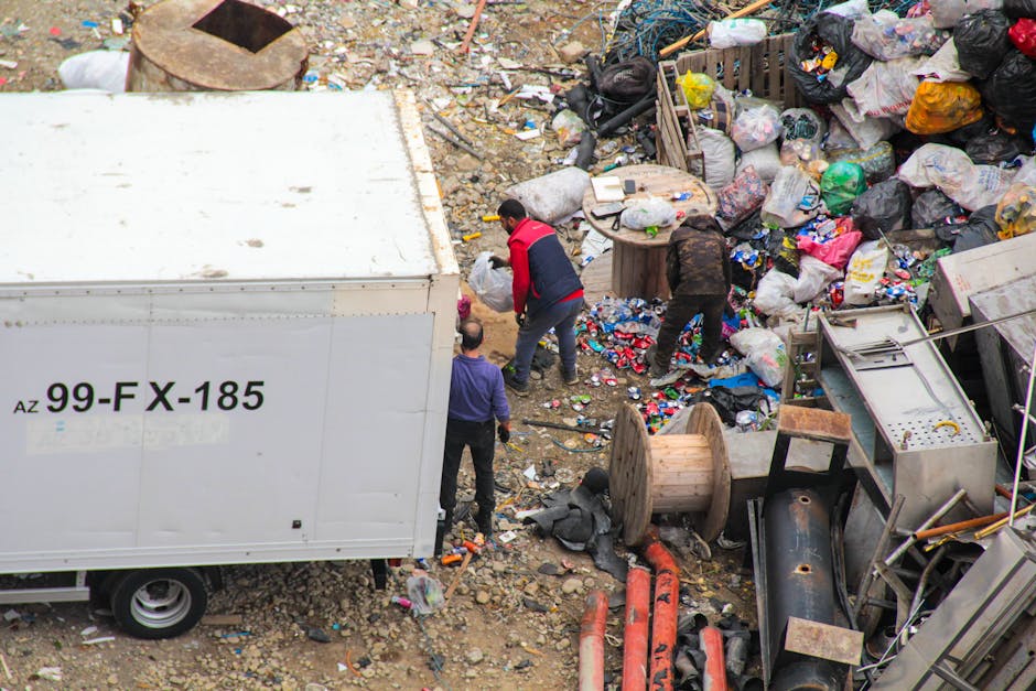An outdoor rubbish collection scene showing two individuals sorting through a large pile of mixed waste materials, including plastic bags, cardboard, and miscellaneous debris, on an uneven, gravelly ground surrounded by discarded items, wooden spools, and industrial equipment. A white waste container with the code '99-FX-185' is positioned on the left side of the image, partially obscuring part of the scene. One person is dressed in dark clothing with a red jacket, bending over to pick up waste, while another in a purple top observes nearby. The environment appears cluttered and chaotic, indicative of an on-site clearance or private waste collection process typically associated with alternative rubbish removal services such as those offered by Rubbish Removal Brixton. The scene is illuminated by natural daylight, emphasizing the variety of materials and textures in the waste, including plastic, metal, and wood, highlighting the practical aspect of the rubbish removal process with an emphasis on thorough sorting and disposal.