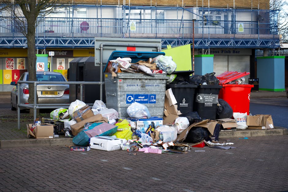 A weathered grey shopping trolley filled with assorted waste materials stands on a narrow, grimy back alleyway amid urban surroundings. Behind the trolley, stacked cardboard boxes, black garbage bags, and other refuse are piled against a dilapidated wall, creating an overflowing scene of rubbish. The alley is flanked by graffiti-covered walls on the right and a rusted, boarded-up entrance on the left, with a few scattered debris and a small black container on the ground. In the background, leafless tree branches and the upper facades of brick buildings are visible, adding an industrial and neglected atmosphere to the scene. The environment appears dimly lit, emphasizing the cluttered and unkempt nature of the space. This scene exemplifies an area where private rubbish collection services, such as those offered by Rubbish Removal Brixton, may assist in clearing excess waste and improving urban cleanliness, especially in alleyways prone to unmanaged debris accumulation.